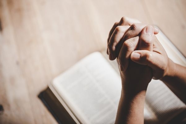 an image of praying hands over a book in the background on a hardwood surface.