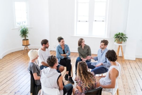 an image of a group of eight individuals gathered in a circle having a discussion, wood floors and white background with window in the background.