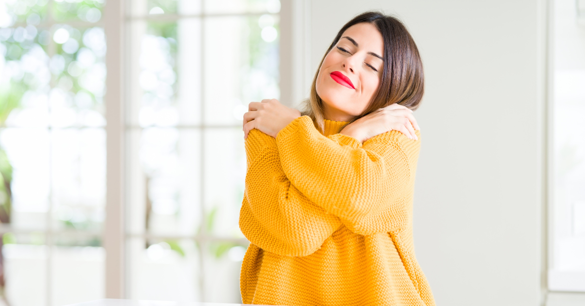 an image of a woman wearing a yellow sweater giving herself a hug on a white background.